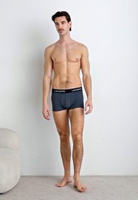 Boxer-briefs gris avec une ceinture noire affichant "LACOSTE." Le mannequin a une silhouette mince et se tient pieds nus dans un environnement minimaliste.