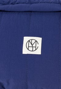 Valkoinen kangasmerkki, jossa on musta ympyräinen monogrammi keskellä sinistä päällystettyä kangasta, jossa on näkyvät ompeleet.