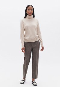 Beiger Rollkragenpullover kombiniert mit karierter Hose in Brauntönen und Orange. Schwarze Schuhe mit weißen Akzenten runden das Outfit ab.