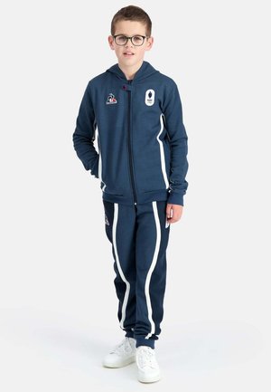 Chándal azul marino con rayas blancas laterales, sudadera con capucha con cremallera y puños elásticos, combinado con zapatillas blancas. Con logotipos en el pecho y los pantalones.