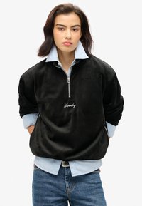 Pullover in velluto a coste nero con colletto a zip, decorato con logo ricamato bianco, indossato sopra una camicia azzurra a bottoni e jeans blu.