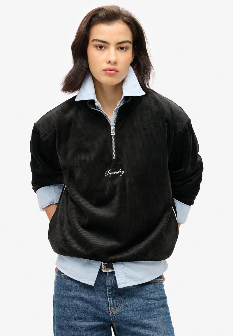 Pullover in velluto a coste nero con colletto a zip, decorato con logo ricamato bianco, indossato sopra una camicia azzurra a bottoni e jeans blu.