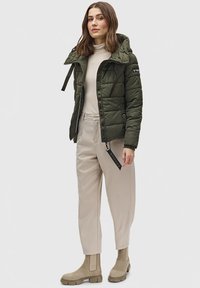 Olijfgroene puffer jas met een capuchon, ritssluiting, zijzakken, zwarte hardware-accenten, gedragen over een lichtbeige turtleneck en broek.