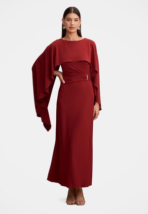Femme portant une longue robe rouge avec de larges manches drapées, une taille cintrée avec une broche, debout avec une main sur la hanche et une expression neutre.