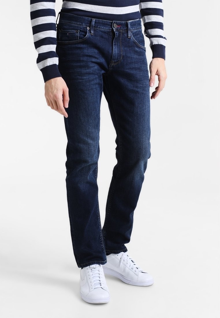 En man som bär mörkblå jeans, vita sneakers och en långärmad tröja med navy- och vita ränder, står mot en vit bakgrund.