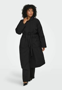 Melnā pildītā jaka ar jostu viduklī, garām piedurknēm un divām priekšējām kabatām. Gluda tekstūra, līdz ceļiem garš un ar apkakles izgriezumu.