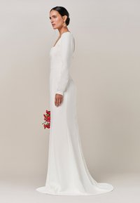 Abito bianco a maniche lunghe con corpino aderente e gonna fluida. Presenta un colletto a quadrato, bottoni decorativi e un mazzo di fiori in mano.