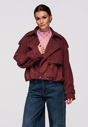 Femme portant une veste oversize bordeaux, un haut rose avec des cœurs rouges, et un jean taille haute bleu foncé, posant devant un fond gris clair.