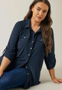 Camisa de botones azul marino con dos bolsillos en el pecho, mangas largas remangadas hasta los codos, fabricada con una tela suave y lisa. Acentos de botones plateados.