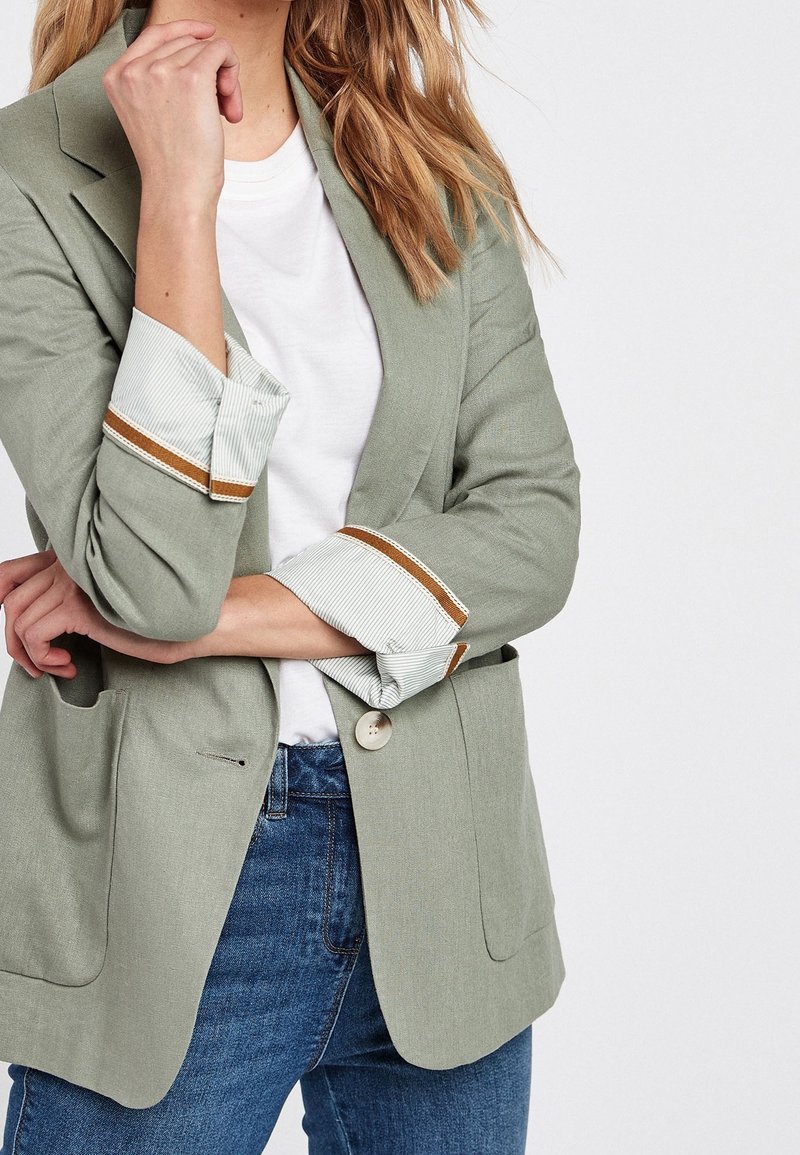 Blazer de lino verde claro con un cierre de botón único, bolsillos parche y puños a rayas con un detalle en marrón. Llevado sobre una camiseta blanca y jeans.