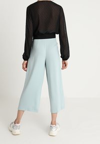 Haut transparent noir à manches longues avec des pois, associé à un pantalon large bleu clair. Le mannequin porte des chaussures de sport argentées.