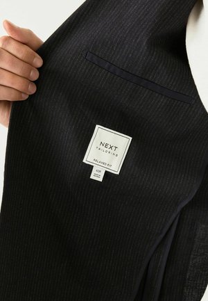 Hand holds a dark striped suit jacket revealing a tag that reads "NEXT TAILORING RELAXED FIT 40R MADE IN VIETNAM."