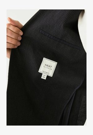 Ręka trzyma ciemną, pasiastą marynarkę, odsłaniając metkę z napisem "NEXT TAILORING RELAXED FIT 40R MADE IN VIETNAM."