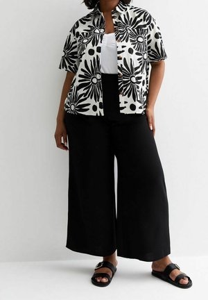 Woman wearing short-sleeve black and white floral button-up shirt, white top, wide black pants, and black sandals with buckle detail.