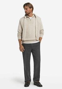 Beige gestrickter Pullover mit Kapuze, über einem weißen Hemd getragen. Graue maßgeschneiderte Hose und schwarze Schuhe mit braunen Schnürsenkeln runden das Outfit ab.