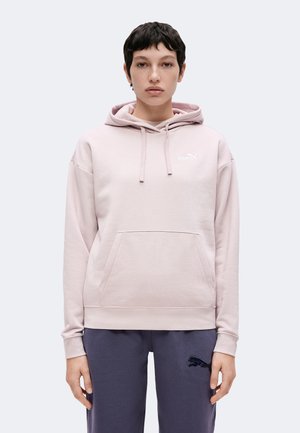 Osoba z krótkimi ciemnymi włosami, ubrana w jasnoróżową bluzę z kapturem Puma oraz ciemnoszare spodnie z logo Puma, stojąca na jednolitym tle.