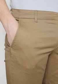 Béžové bavlněné kalhoty mají slim fit, postranní kapsy a hladkou strukturu, s klasickým pasem pro opasek.