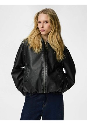 Blondínka v čiernej koženej bunde na zips a tmavomodrých džínsoch, stojaca s rukami v kapsách bundy proti jednoduchému pozadiu.