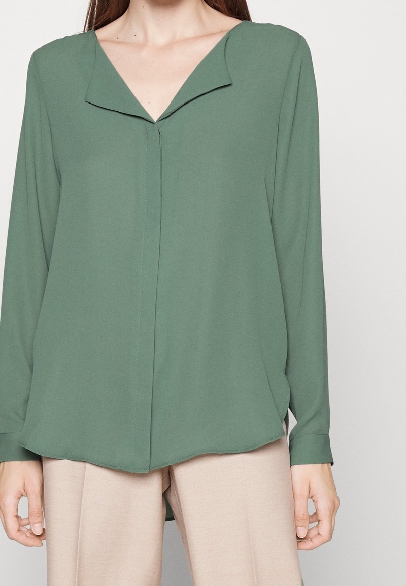 Blouse à manches longues en v en tissu vert muted avec une texture lisse, présentant une couture frontale discrète et un ourlet arrondi.