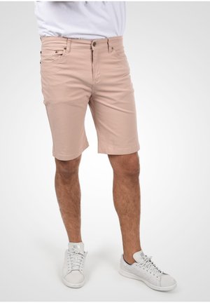 Mann trägt beige kniehohe Shorts, weiße Turnschuhe und ein weißes Hemd, steht vor einem schlichten hellen Hintergrund.