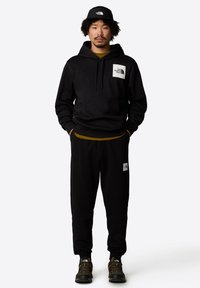 Czarna bluza z kapturem z kieszenią kangurzą i naszywką z logo, w połączeniu z pasującymi czarnymi dresami i czarnymi sneakersami z żółtymi akcentami.