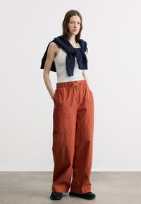 Jeune femme debout, les mains dans les poches, portant un débardeur blanc, un pantalon large couleur rouille, des baskets noires et un sweat-shirt bleu marine posé sur les épaules.