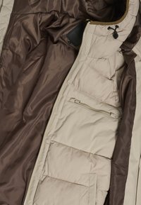 Beige pufferjacka med brunt satinfoder, har en hel dragkedja, fickor och en dragsko vid huvan. Kviltat mönster.