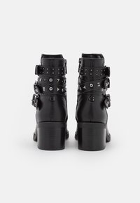 Botas de cuero negras hasta el tobillo con un tacón ancho, que presentan múltiples correas con tachuelas plateadas y adornos de hebilla. Textura suave y diseño detallado.