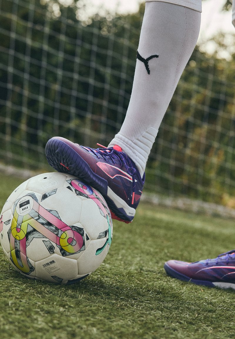 Ljubičaste i ružičaste sportske cipele s teksturiranim potplatom odmaraju na šarenoj nogometnoj lopti. Pozadina s travom i mreža gola su vidljive.