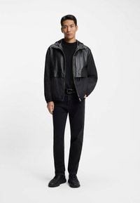 Schwarze Bomberjacke mit glänzendem Lederobereinsatz und mattem unteren Teil. Kombiniert mit schwarzen Jeans und schwarzen Sneakers.