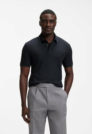 Mann trägt ein schwarzes Poloshirt mit kurzen Ärmeln und hellgraue Hose, steht mit einer Hand in der Tasche vor einem einfarbigen Hintergrund.