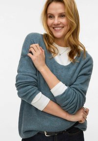 Gaiši zils ribots džemperis ar plašu kakla izgriezumu un garām piedurknēm, pār muguras valkāts virs baltas blūzes. Gluda struktūra, brīvs piegriezums un ikdienišķs stils.