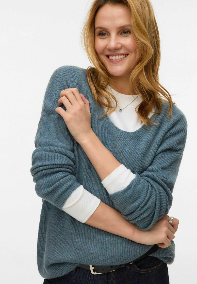 Gaiši zils ribots džemperis ar plašu kakla izgriezumu un garām piedurknēm, pār muguras valkāts virs baltas blūzes. Gluda struktūra, brīvs piegriezums un ikdienišķs stils.