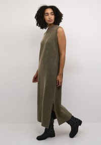 Robe sans manches en tricot vert olive avec des fentes sur les côtés, texture côtelée et silhouette droite, associée à des bottes noires mi-mollet.