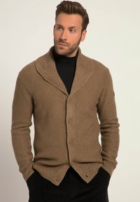 Brunstrikket cardigan med sjalskrave og knaplukning. Har tekstureret stof og buet kant. Bæres over en sort turtleneck.