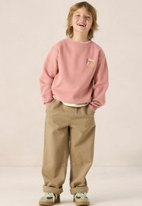 Roze sweatshirt met een logopatch, losse beige broeken en groen-witte sneakers. De stof lijkt zacht met een relaxte pasvorm.