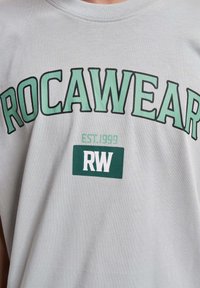 Vaaleanharmaa t-paita, jossa on vihreät kirjaimet "ROCAWEAR" ja "EST.1999" vihreän suorakulmion yläpuolella, jossa on valkoiset "RW"-kirjaimet. Pehmeä kankaan tekstuuri.