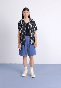 Chemise noire à manches courtes avec des motifs yin-yang blancs, assortie à un short en denim bleu et des baskets montantes beiges, se tenant sur une surface bleu clair.