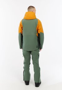 Groene en oranje waterdichte jas met capuchon, voorzien van een aangesloten design en verstelbare manchetten. De broek is kleur afgestemd en heeft ritsen bij de enkels.