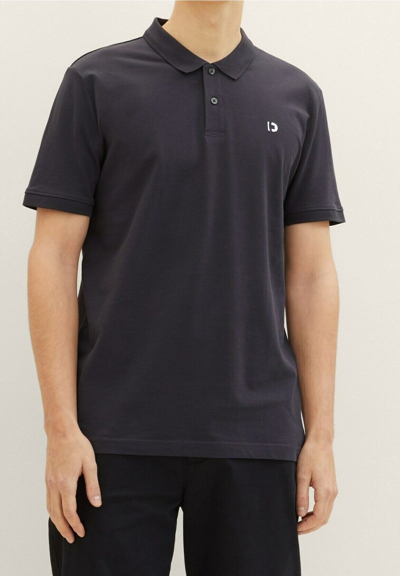 Homme portant un polo sombre uni avec deux boutons et un petit logo blanc sur le côté gauche de la poitrine, se tenant devant un fond clair.