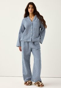 Kobieta nosząca niebiesko-białą w kratę piżamę z długimi rękawami i zapinaną na guziki bluzką, w połączeniu z futrzanymi kapciami w zwierzęcy wzór.