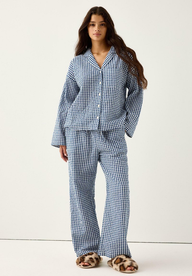 Kobieta nosząca niebiesko-białą w kratę piżamę z długimi rękawami i zapinaną na guziki bluzką, w połączeniu z futrzanymi kapciami w zwierzęcy wzór.