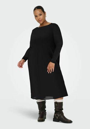 Robe noire à manches longues avec un col bateau, au design fluide et texture lisse. Portée avec des bottes marron à hauteur de genou et des chaussettes blanches.