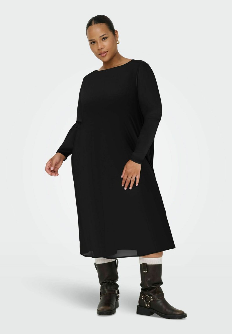 Robe noire à manches longues avec un col bateau, au design fluide et texture lisse. Portée avec des bottes marron à hauteur de genou et des chaussettes blanches.