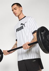 Hvid stribet træningstrøje med sorte accenter og Puma-logo, parret med sorte shorts. Individ løfter en vægtstang med synlige tatoveringer.