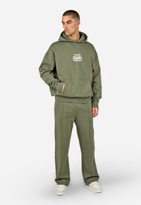 Felpa con cappuccio verde oliva e pantaloni della tuta coordinati, entrambi realizzati in tessuto morbido. La felpa presenta una tasca frontale e un logo sul petto.