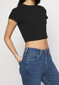 Mujer vistiendo una camiseta negra corta y unos jeans de talle alto azul, con la mano derecha en el bolsillo, frente a un fondo liso.