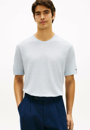 Ung mand iført en lyseblå kortærmet striktrøje, stoppet ned i mørkeblå bukser med læg, stående med hænderne i lommerne.