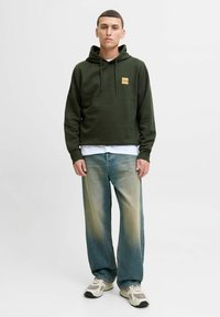 Dark green hoodie with a front logo patch, paired with light blue wide-leg jeans and beige sneakers, set against a plain background.