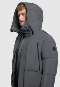 Muškarac nosi zimski kaput od sive podstavljene tkanine s prevelikim kapuljačom, zatvorenim džepom na prsima i podešavanjem uzicom.